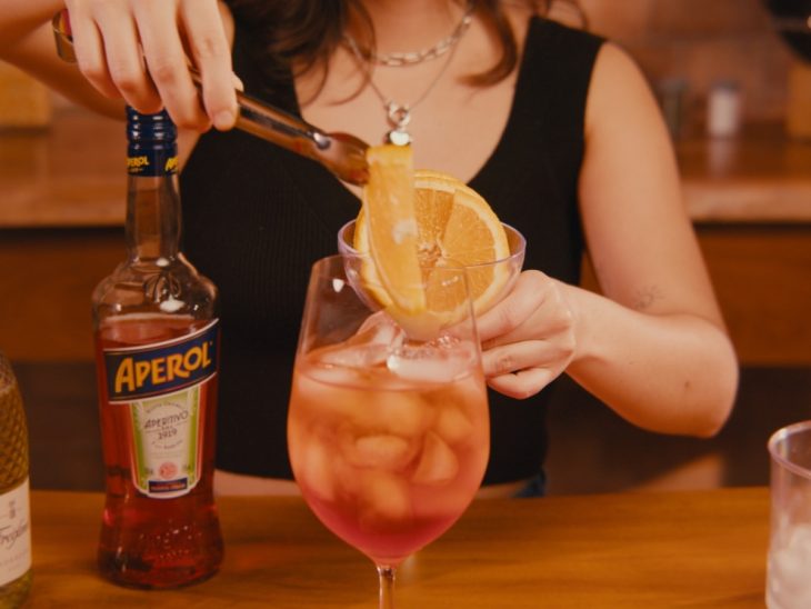 Uma taça contendo aperol spritz sendo decorada com rodelas de laranja.