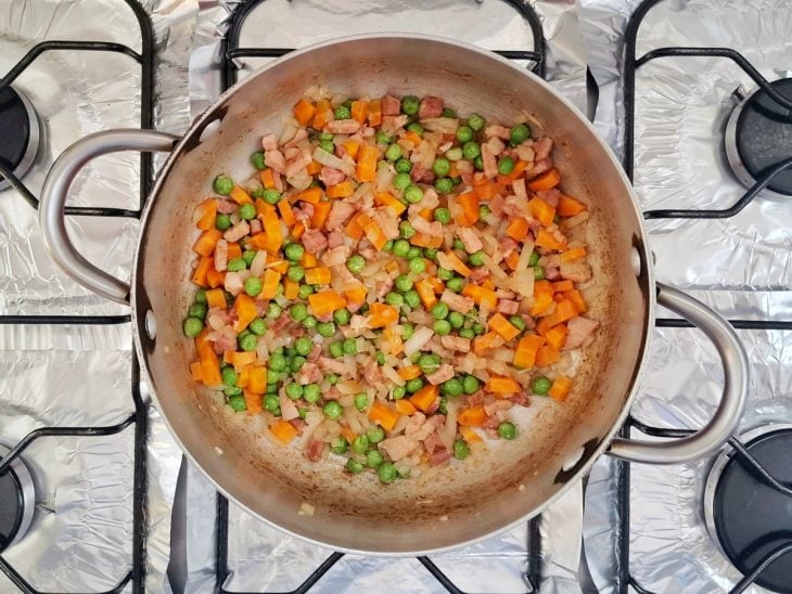 Panela no fogo com manteiga, alho, cebola, ervilha e cenoura cozidos.