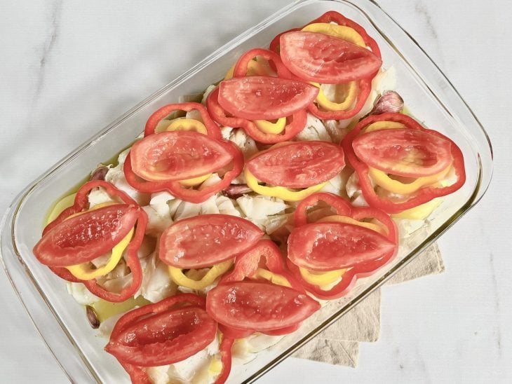 Um refratário com camadas de tomate, pimentão, bacalhau e azeite ao fundo.