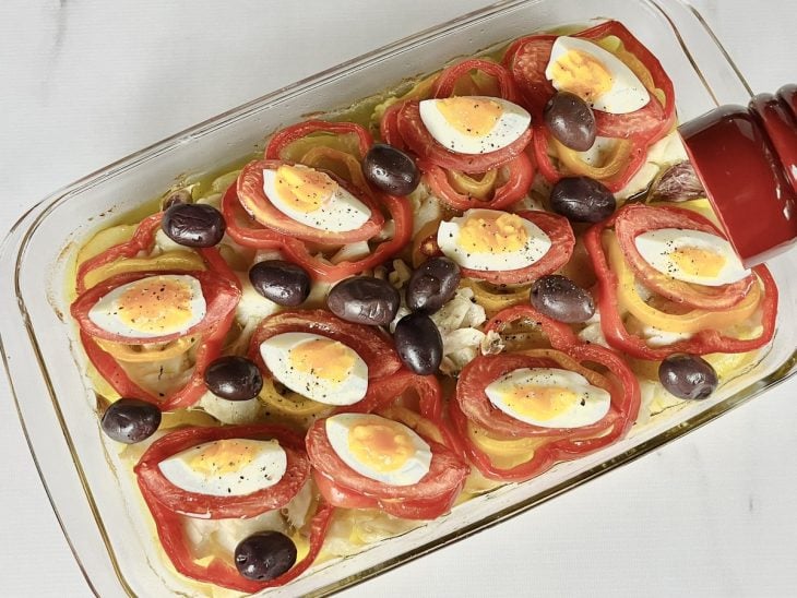 Um refratário com camadas de ovos, azeitonas, tomate, pimentão, bacalhau e azeite ao fundo.