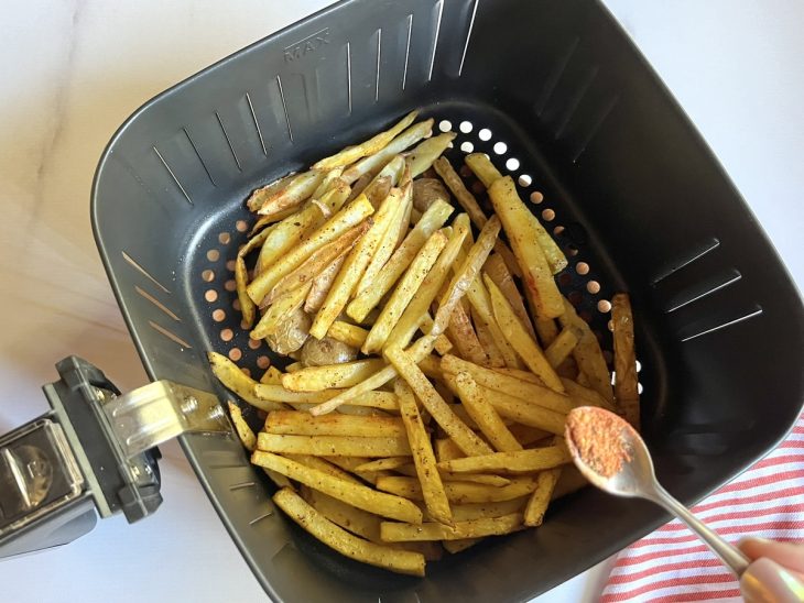 Uma cesta de airfryer com as batatas fritando e a adição de temperos.