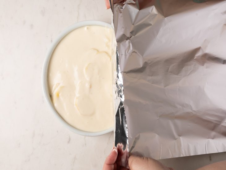 Um refratário com as batatas e molho branco sendo coberto com papel-alumínio.