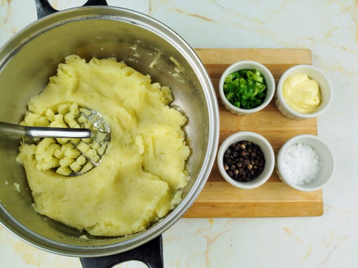 Batata sendo amassada por um amassador de batatas em uma tigela com potinhos com manteiga, pimenta-do-reino, sal e a cebolinha picada ao lado.