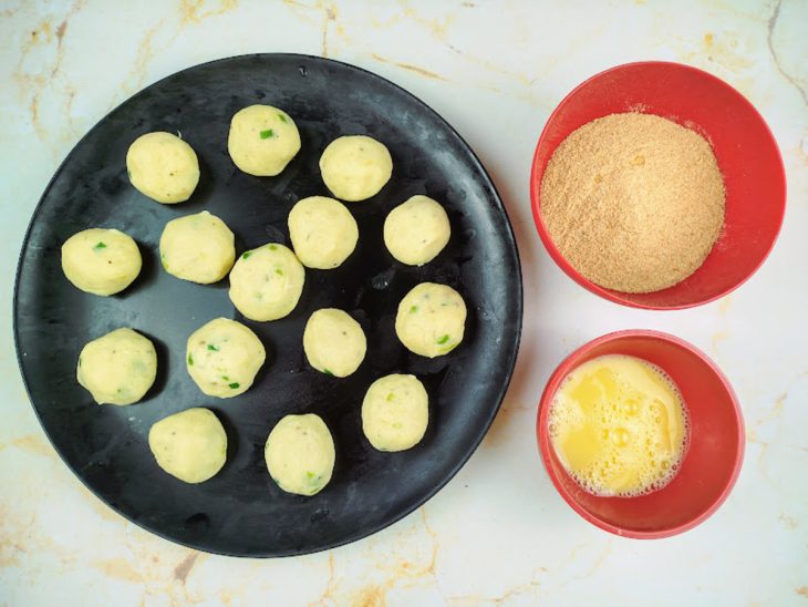 Bolinhas de massa de batata em um prato com dois potes ao lado com farinha de rosca e ovo batido.