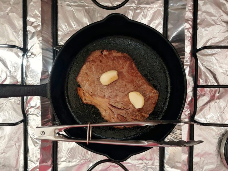 Uma frigideira selando um bife ancho com dentes de alho.