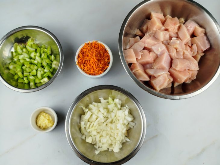 Frango em cubos, cebola picada, alho amassado, cenoura ralada e salsão cortado separados em recipientes.