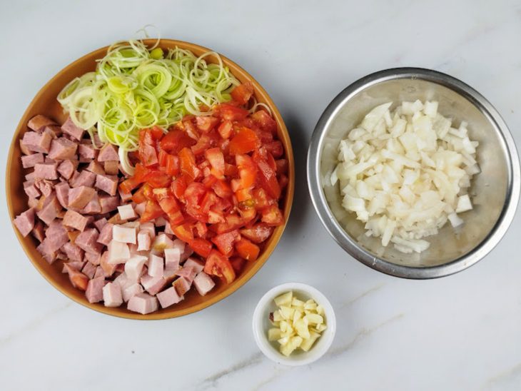 Calabresa, bacon, tomate, alho-poró em uma tigela de cerâmica. Cebola picada em um recipiente de alumínio e alho em um pequeno pote.