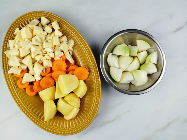 Batatas descascadas e picadas em um prato com a cenoura e o aipim também picados. Cebola picada em um recipiente ao lado.