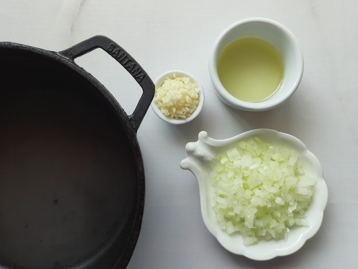 Sobre uma bancada, azeite, alho e cebola em recipientes e uma panela ao lado