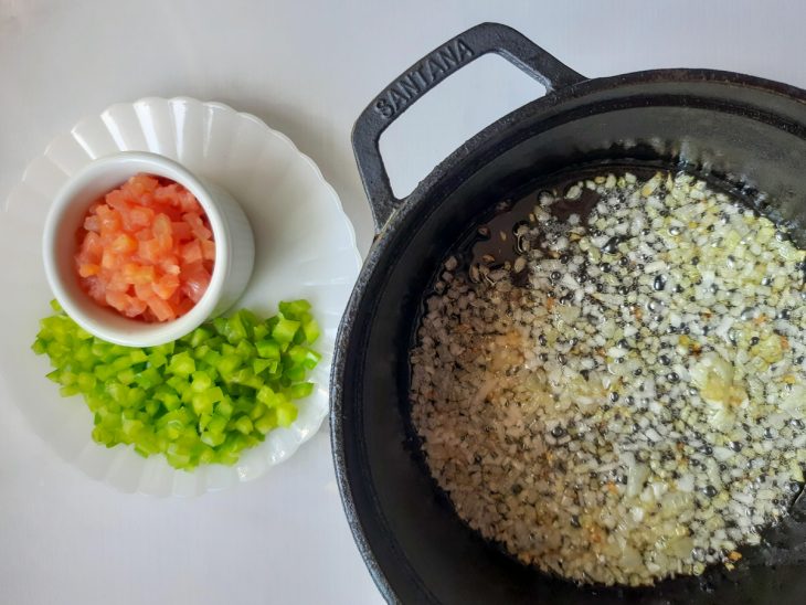 Alho e cebola refogados em uma panela. Tomate e pimentão picados em recipientes ao lado