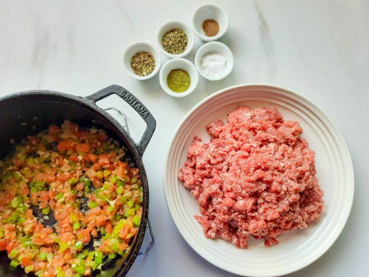 Panela com ingredientes refogados, carne moída crua em um prato e temperos variados em recipientes ao lado.