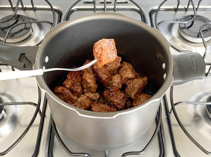 Pedaço de carne de panela espetado em um garfo e demais pedaços na panela.