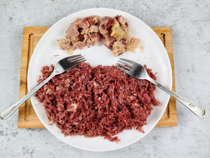 Carne sendo desfiada e separada da gordura por um garfo em um prato.