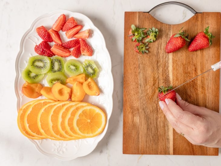 Um prato com laranja, pêssego, kiwi e morango picados, e uma tábua com morangos sendo cortados.