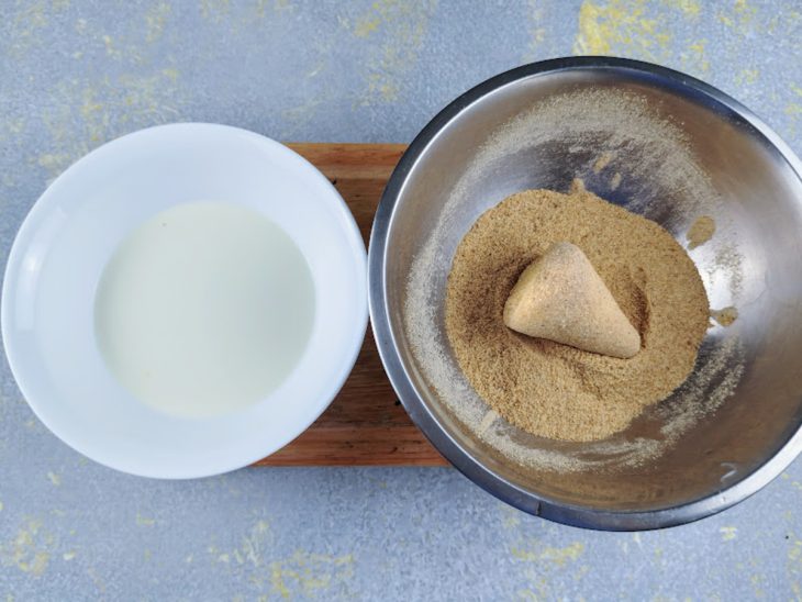 Tábua com duas tigelas, de leite e de farinha de rosca e uma coxinha sendo empanada.