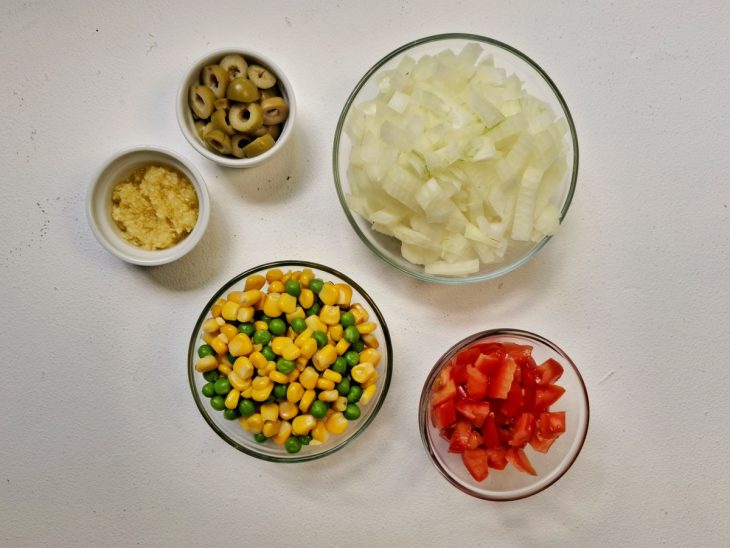 Alho, cebola, tomate, azeitona, milho e ervilha já cortados e separados em recipientes diferentes.