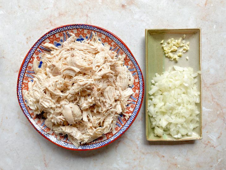 Um prato com o frango desfiado e outro recipiente com alho e cebola picados.