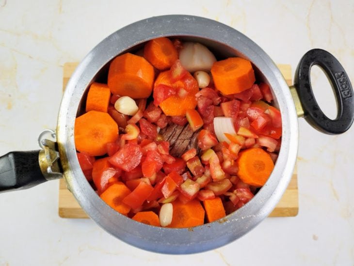 Uma panela de pressão com a carne sobre a camada de legumes e coberta com mais uma camada.