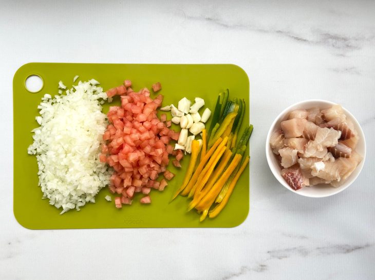 Alho, cebola, pimentão e tomate em cortes sobre uma tábua, e ao lado, recipiente com pintado em cubos.