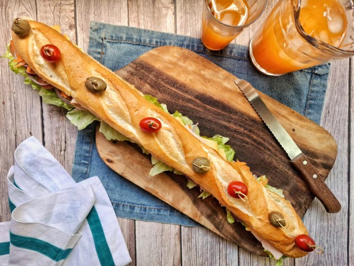 Pão decorado com tomate e azeitona sobre uma bancada com utensílios de cozinha e recipientes com suco pronto para servir.