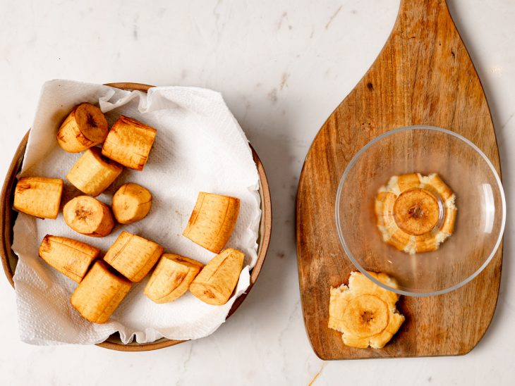 Uma tábua com as bananas fritas sendo amassadas.