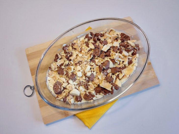 Um refratário forrado com creme de chocolate e coberto com bolachas, creme branco e bombons picados.
