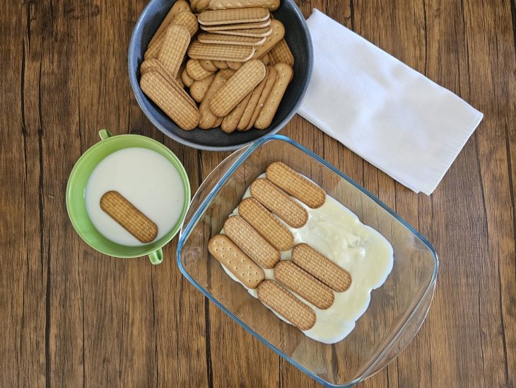 Um refratário forrado com uma camada de creme branco sendo coberto por bolachas.