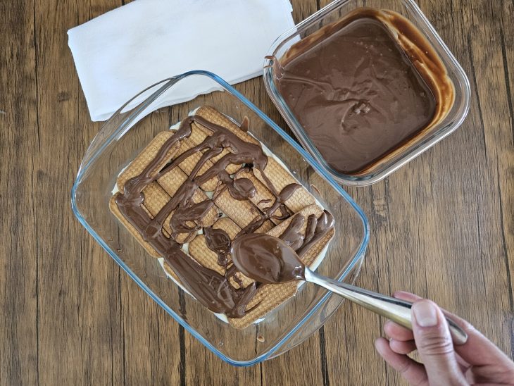 Um refratário forrado com uma camada de creme branco sendo coberto por bolachas e ganache.