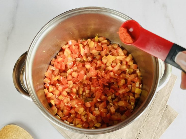 Uma panela com azeite, cebola, alho, pimentões, tomate, pimenta-de-cheiro e colorau.