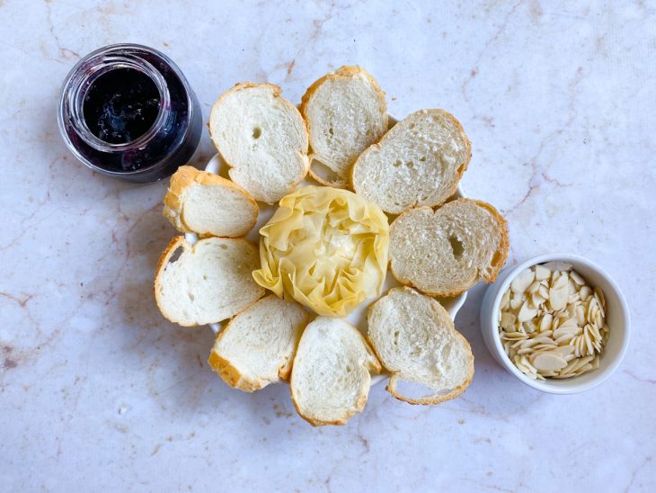 trouxinha de queijo brie com massa filo já assada em uma travessa com torradas e um pote de geleia ao lado e um pote com lâminas de amêndoas.