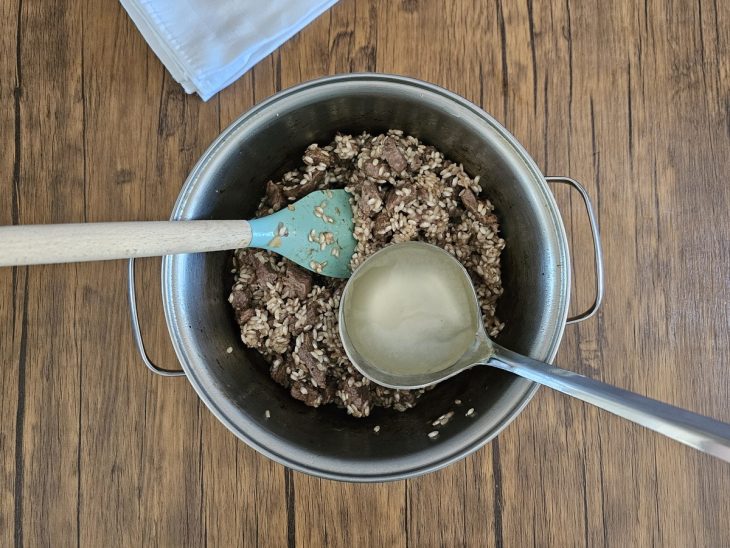 mistura de carne com arroz e vinho sendo mexida por uma espátula e uma concha de caldo sendo adicionada.