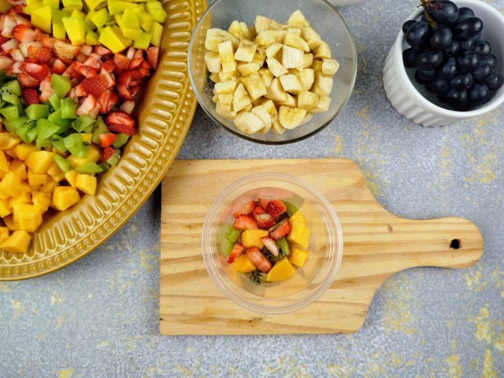 Pote com três tipos de frutas picadas. Tábua com várias frutas picados ao lado.