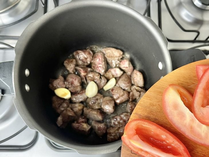 Uma panela de pressão selando pedaços de carnes, com alho e tomate.