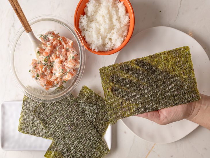 Folha de nori em uma mão e recipientes com salmão e arroz em volta.