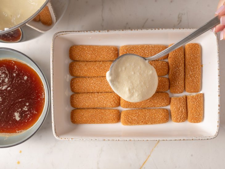 Um refratário com uma camada de bolachas champanhe e uma porção de creme sendo derramada.